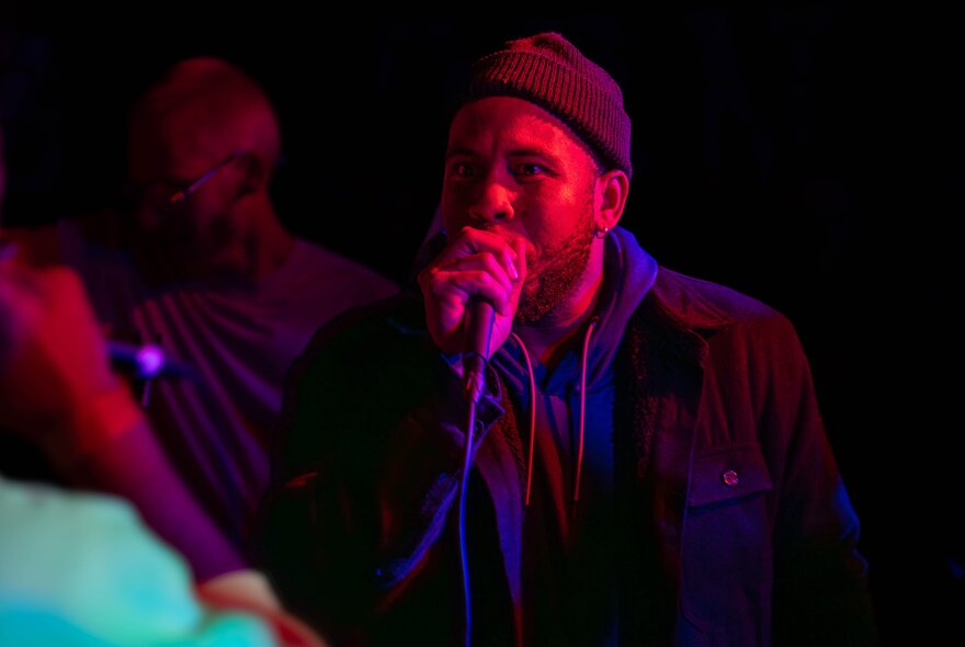 A man in a beanie performs into a microphone in a dark room with a red light on his face. 