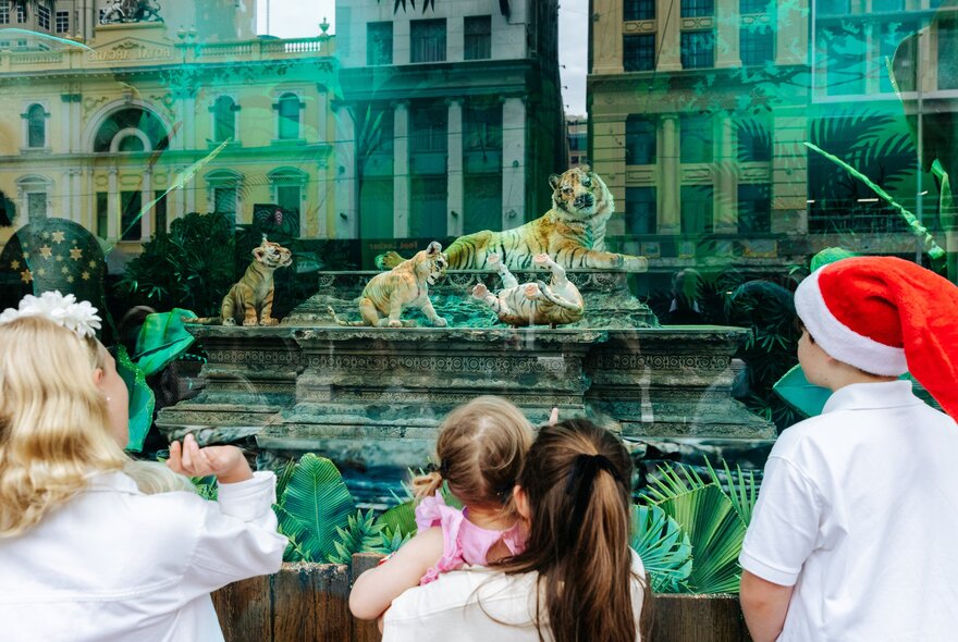 A family looking at the Myer Christmas Windows showing a tiger mum and her cubs, the city buildings in the street behind them reflected in the glass.