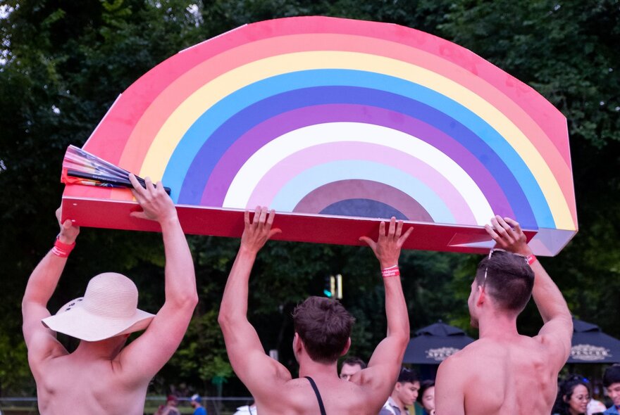 Three people carrying a large rainbow sign in a crowded place.