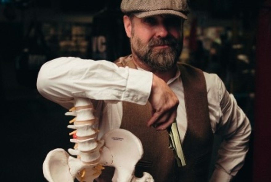 Bearded man wearing a cap and waistcoat, leaning his arm on the headless spine of a skeleton.