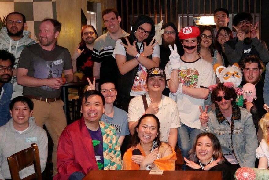 A large group of smiling people, some dressed in cosplay, posing around a table at a trivia night being held in a large room.