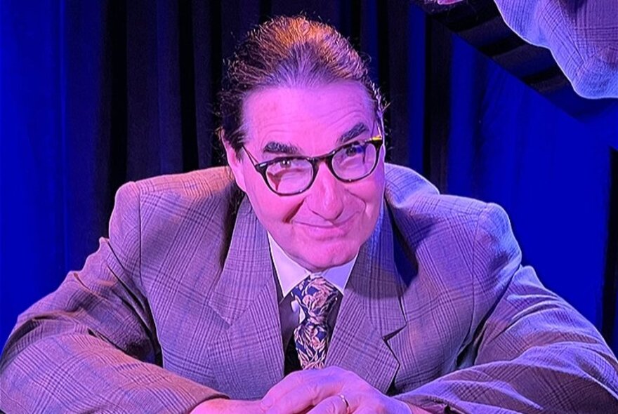 A man dressed as Tom Lehrer with hair slicked back and glasses under a pink / purple light. 