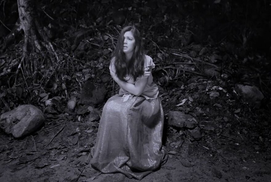 Black and white still from the film Season of the Devil, shows a woman with long, dark hair wearing a long seated on the ground in a dark, wooded area.
