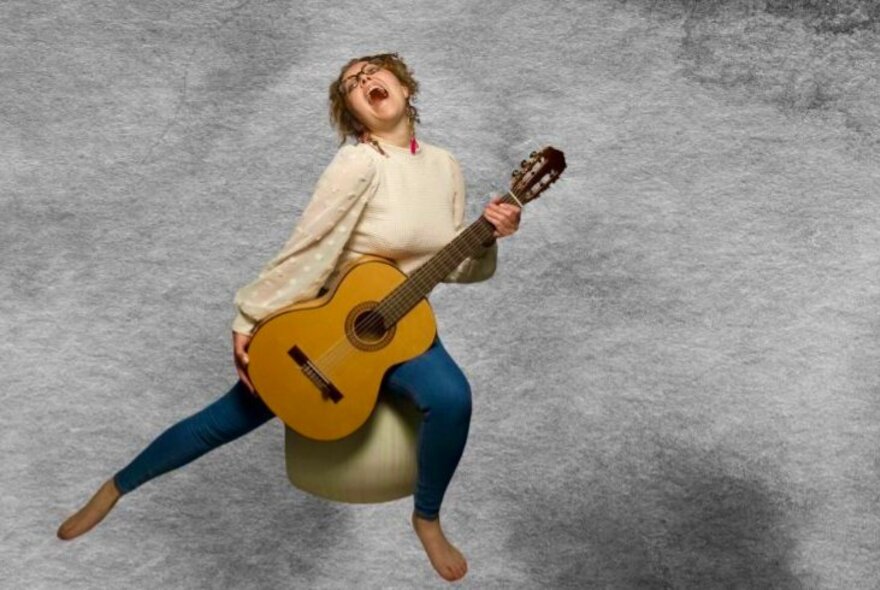 A woman sitting on an exercise ball, singing passionately while playing an acoustic guitar, against a pale grey background.