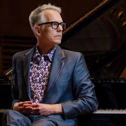 Male pianist wearing a shiny blue suit, resting his hands on his lap while leaning on a piano.