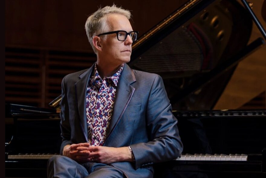 Male pianist wearing a shiny blue suit, resting his hands on his lap while leaning on a piano.