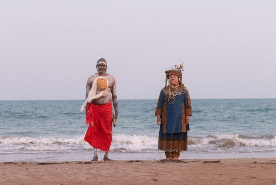 Two people standing upright on sand at the shoreline of a beach, each wearing clothes that represent their country and place.