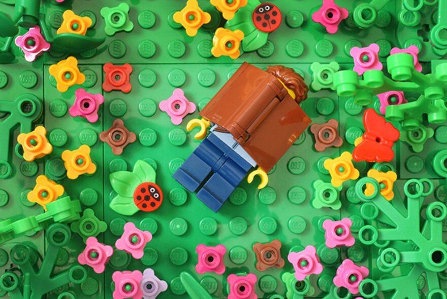 Looking down at a green Lego base board with a Lego figurine lying on their back with a book over their face, and flowers all constructed from Lego pieces.