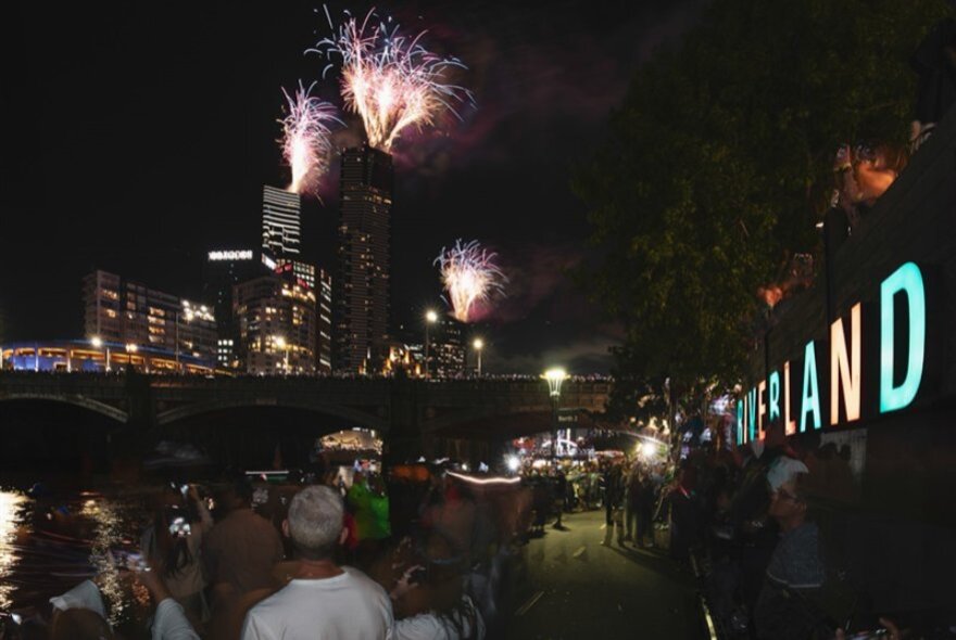 People standing on the wharf outside Riverland Bar at night, watching fireworks over the tops of city buildings.
