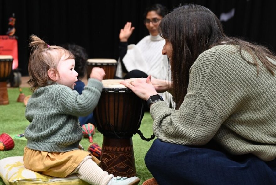 A toddler sitting playing an African drum while an adult woman looks on encouragingly.