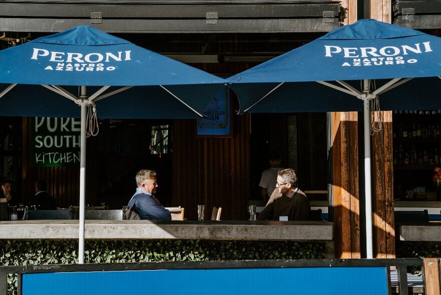 Two men sitting at an outdoor table at a restaurant, with two blue cafe umbrellas in front of them.