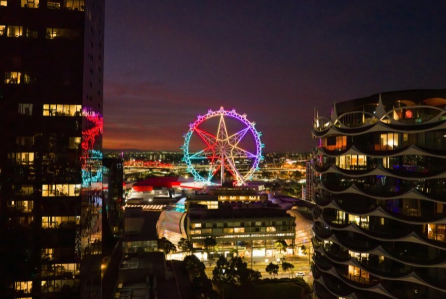 Melbourne Star Observation Wheel at night peeking through other buildings and illuminated in bright colours. 