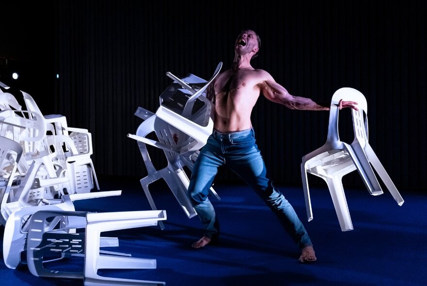 A dancer wearing jeans and with a bare torso, interacting with white plastic chairs on a dance floor stage.