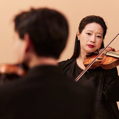 Violinists performing with their instruments, one with their back to the view, the other a woman with black hair.