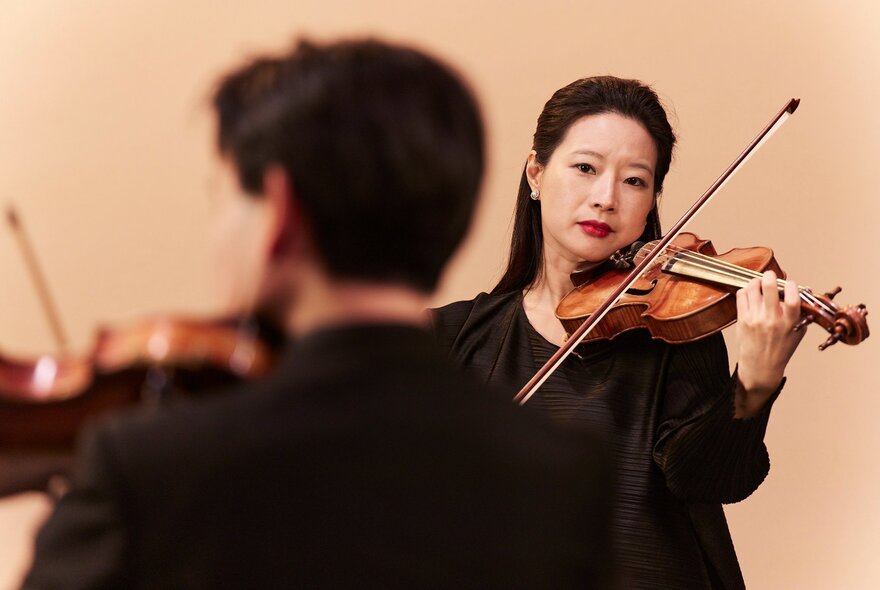 Violinists performing with their instruments, one with their back to the view, the other a woman with black hair.
