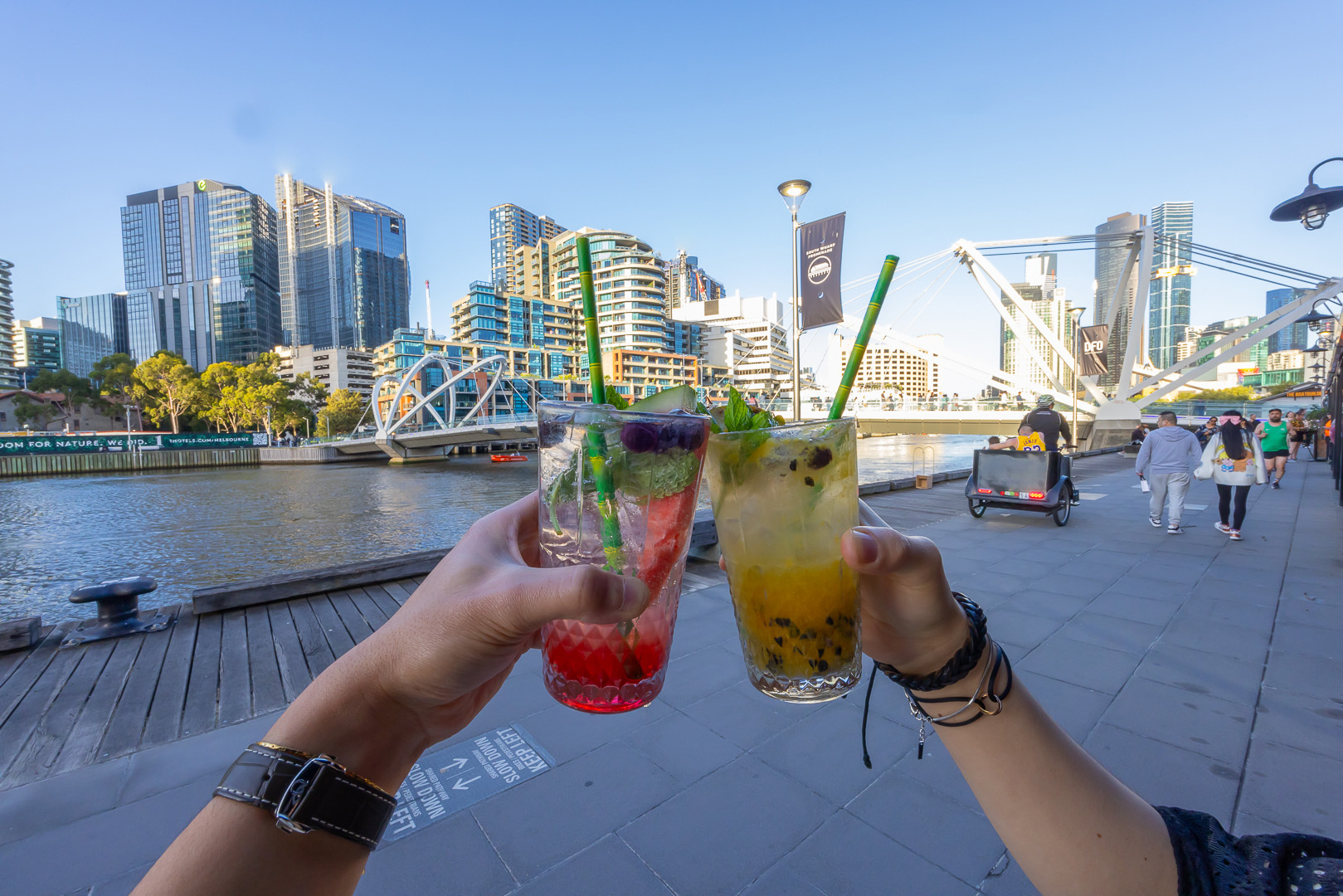 Two people holding colourful drinks in front of the water.