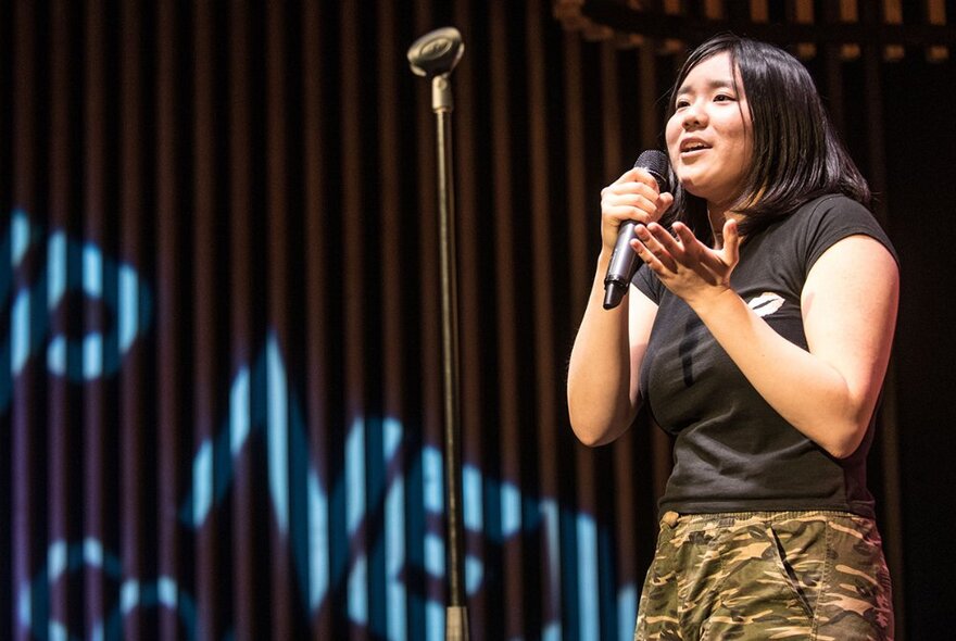 Young teenager holding a microphone in her hand and performing stand up comedy on a stage.