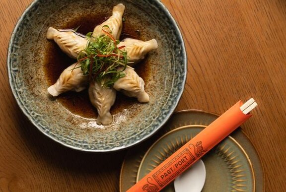 Looking down at a shallow plate of dumplings in a sauce, with chopsticks wrapped in orange paper alongside.