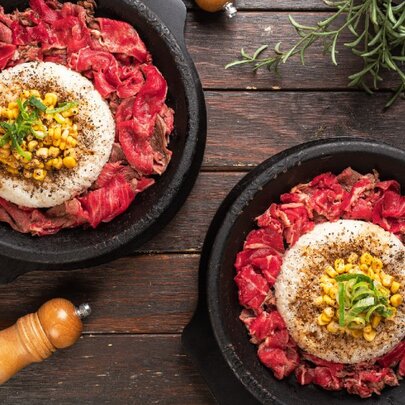 Looking down at two dishes of thinly slice beef with rice and garnishes on top.