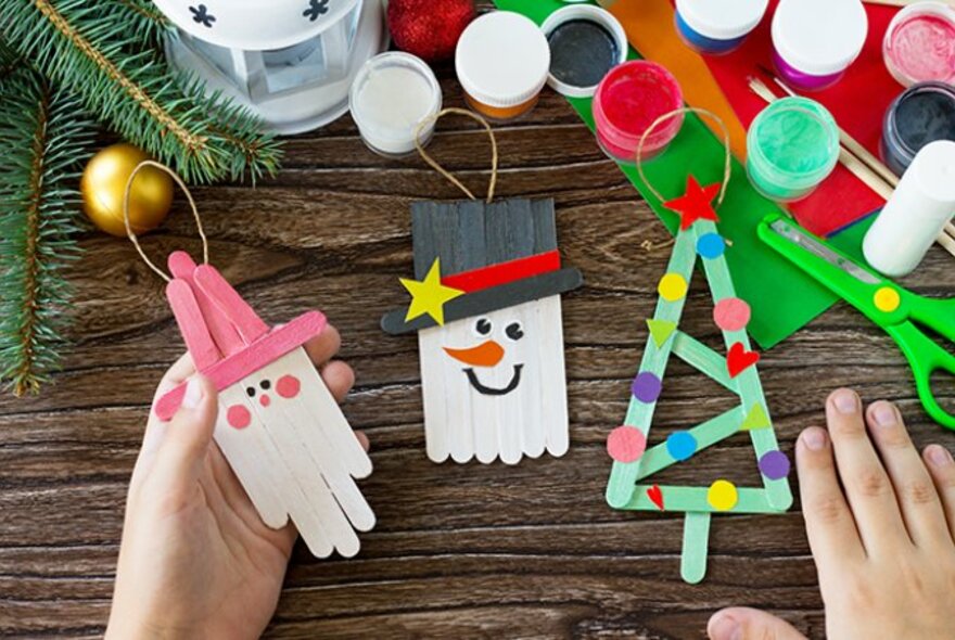 Overhead view of a child's hands at a craft table with small pots of paint, a glue stick, and wooden ice-cream sticks.