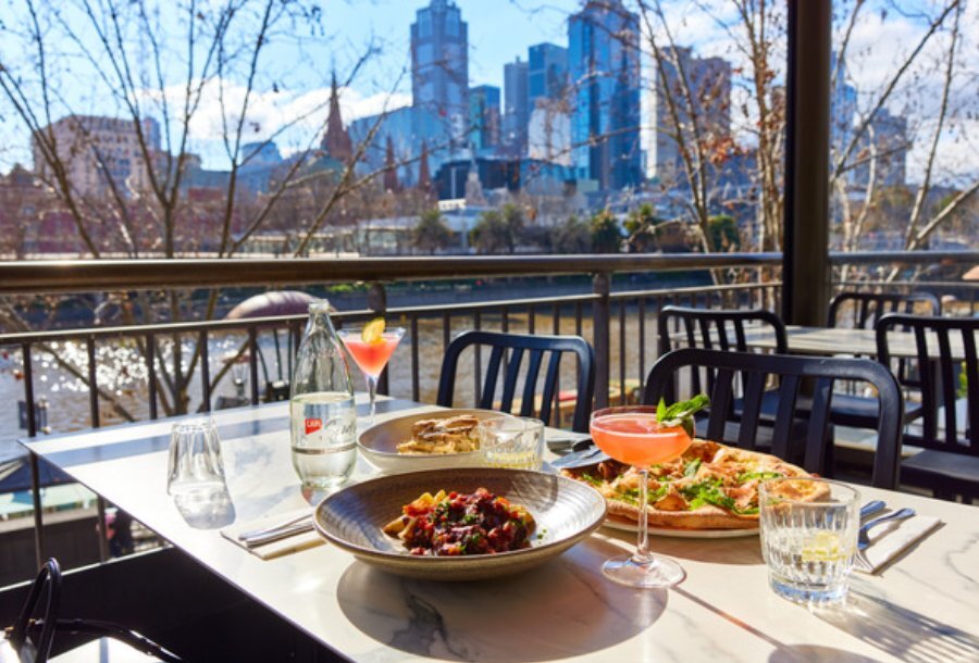 A table with pizza, a bowl of pasta and cocktails seated outside by the Yarra River with the city skyline in the background.