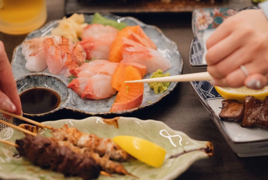 A platter of raw sushi behind a separate platter with yakitori skewers; a person's hand using chopsticks to pick up a piece of sushi.