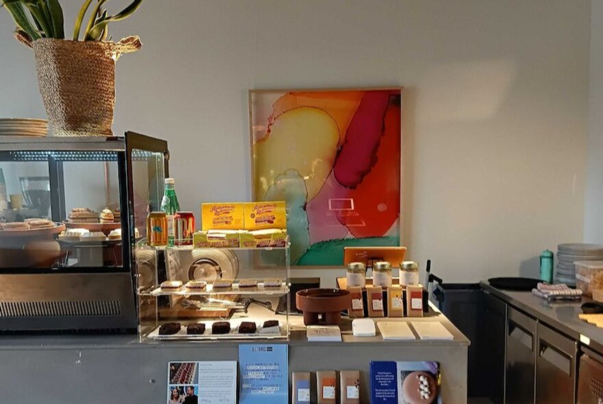 The service counter of a cafe, with food on display behind a glass cabinet, an artwork on the back wall and other products for sale.