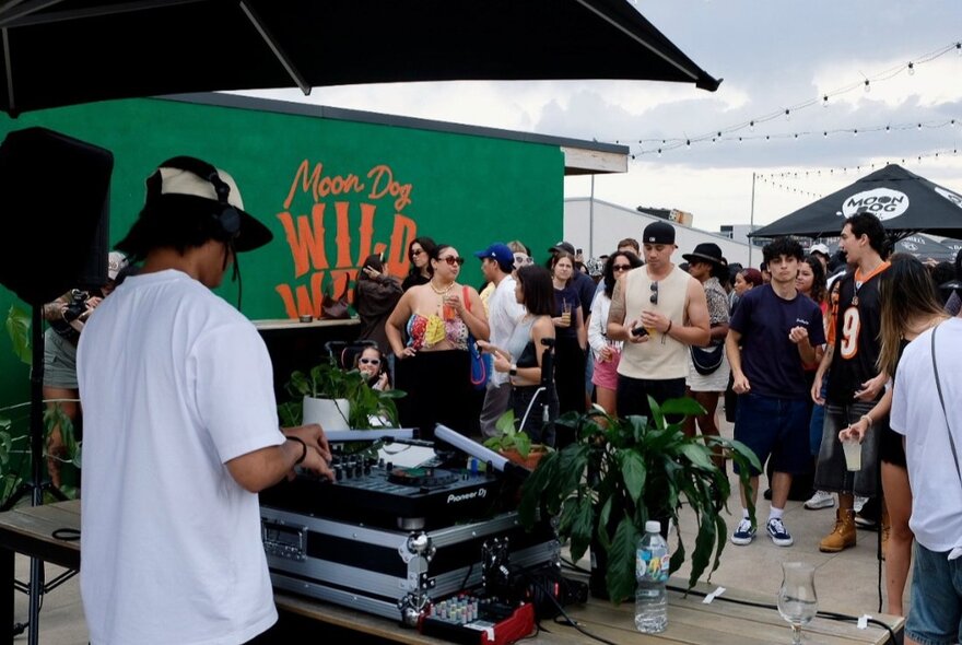 DJ at a music desk at an outdoor rooftop venue, people looking on.