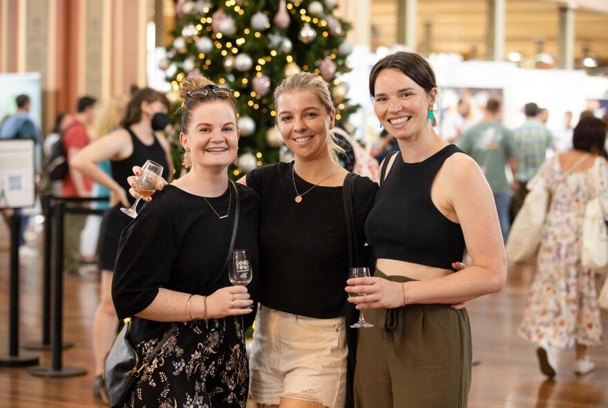 Three people arm in arm holding drinks in front of a Christmas tree.