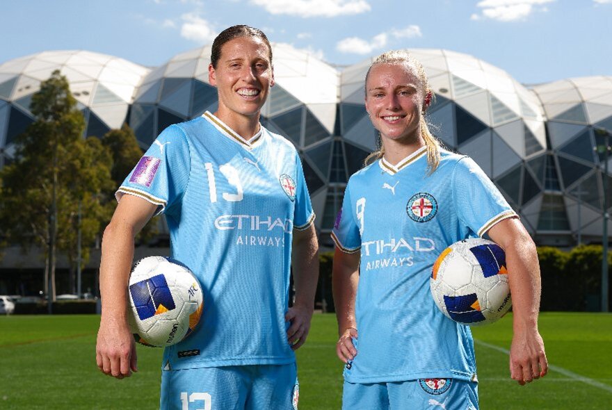 Two players dressed in Melbourne City kit and holding soccer balls in front of AAMI park.