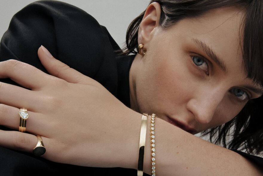 A woman looking over her crossed arms showing two gold bracelets on her wrist and a diamond ring and signet ring on her hand