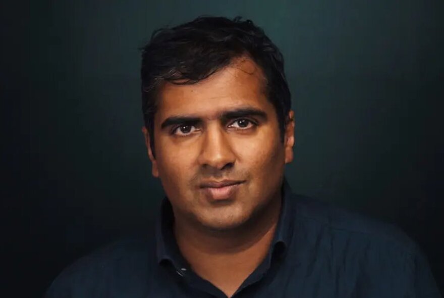 Comedian Sanjay Patel, looking directly at the viewer with a neutral expression, posed in front of a dark background.