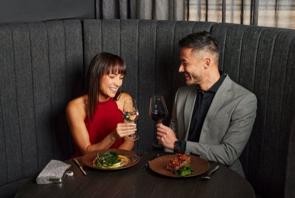 A couple seated in a curved booth at a restaurant, smiling at each other and holding up wine glasses, plates of food before them.