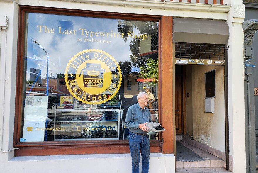 A man holding a typewriter outside the glass shopfront of the last typewriter shop in Melbourne.