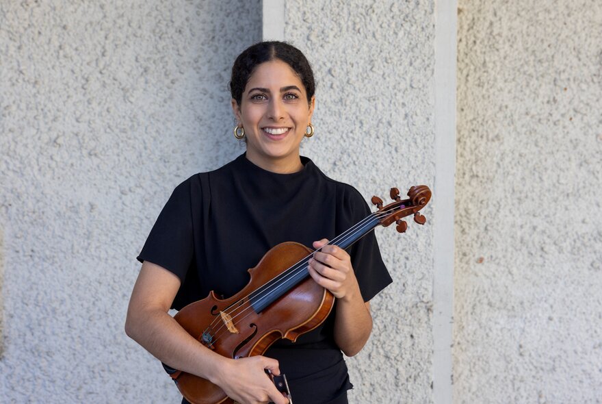 Violinist Lydia Sawires smiling and holding her violin under her arm, standing posed against a white wall.