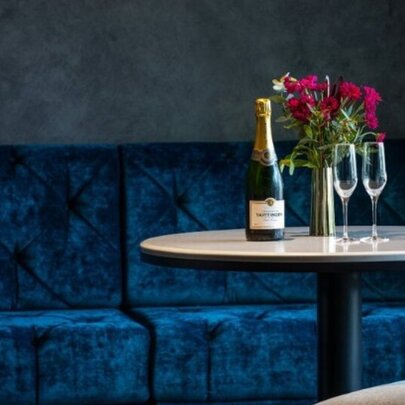 A bottle of sparkling wine on a round table with two glasses and a vase of flowers, in front of deep blue velvet bench seats.