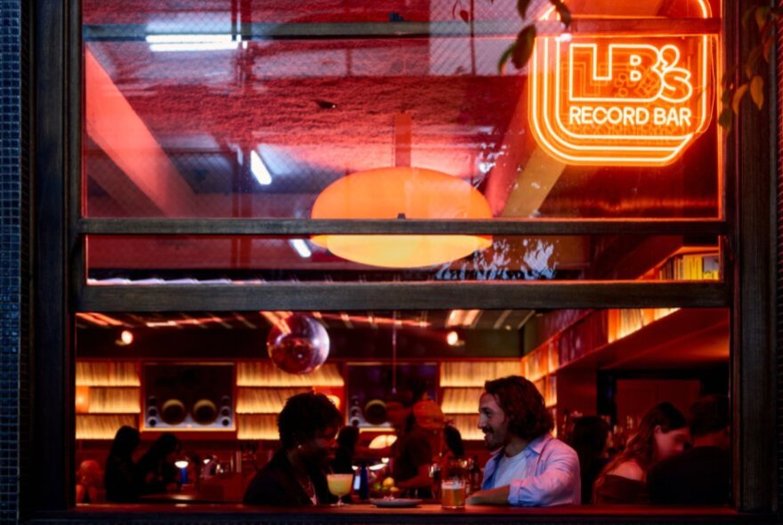 People seated at a window table viewed from outside with backlit shelves of LPs and neon signage.