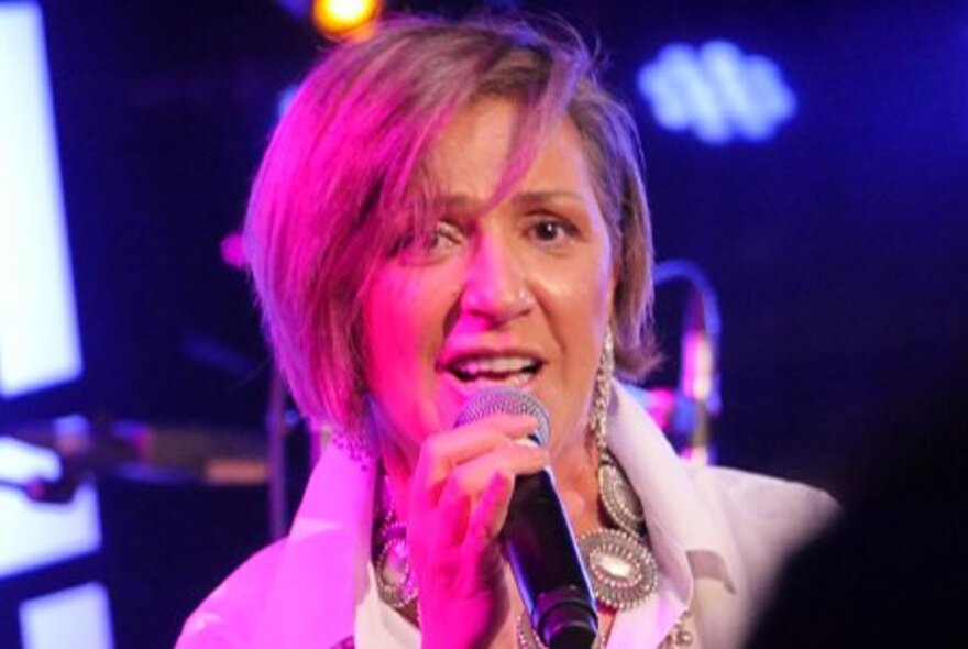 Singer, Noria Letts, singing into a microphone with a pink stage light shining on her short blonde hair.