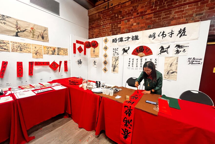 Calligraphy artist working on a red banner with Chinese lettering on a long worktable with craft items, covered with a red cloth.
