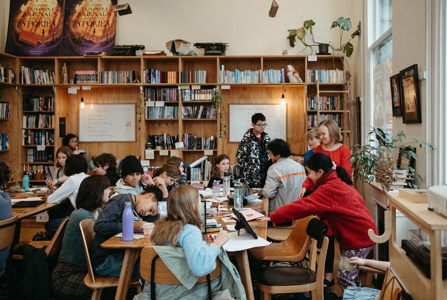 Young writers seated at tables writing in a workshop with papers and books.
