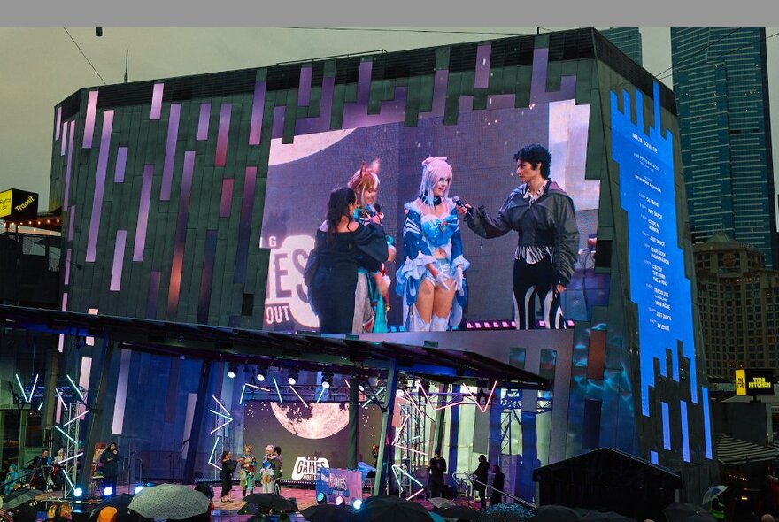 The big screen at Fed Square, showing three people in Cosplay fashions.