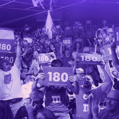 A cheering audience at an indoor darts tournament, some of them holding up signs that have the number '180' printed on them.