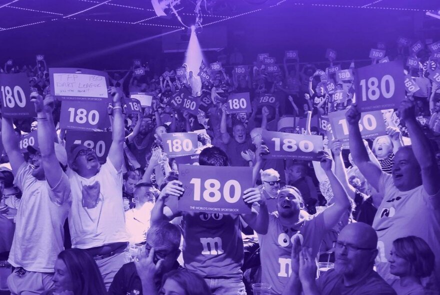 A cheering audience at an indoor darts tournament, some of them holding up signs that have the number '180' printed on them.