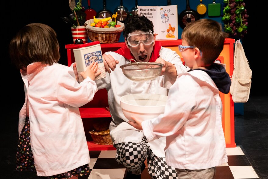 A man wearing safety goggles bends down to cook with two small children - all wearing white shirts. 