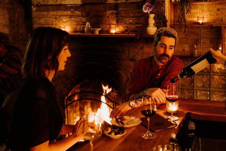 A warm bar in front of a roaring fire with a bartender's hands pouring a glass of red wine for two people at the bar. 