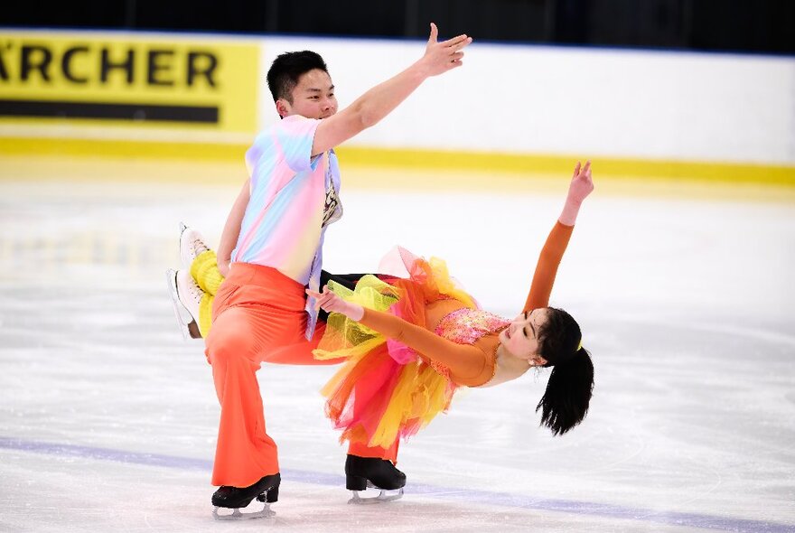 Two young figure skaters performing on the ice, the girl leaning horizontally over the boy's knee.