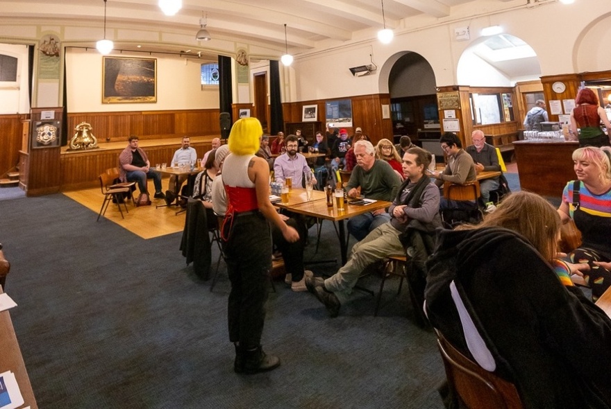 People seated at tables in a bar, looking at a person who is standing in front of them and about to leading them in a song.