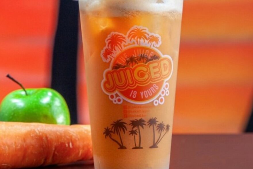 A takeaway plastic cup of fresh orange coloured juice, with branding on the cup, and a green apple and carrot next to it.