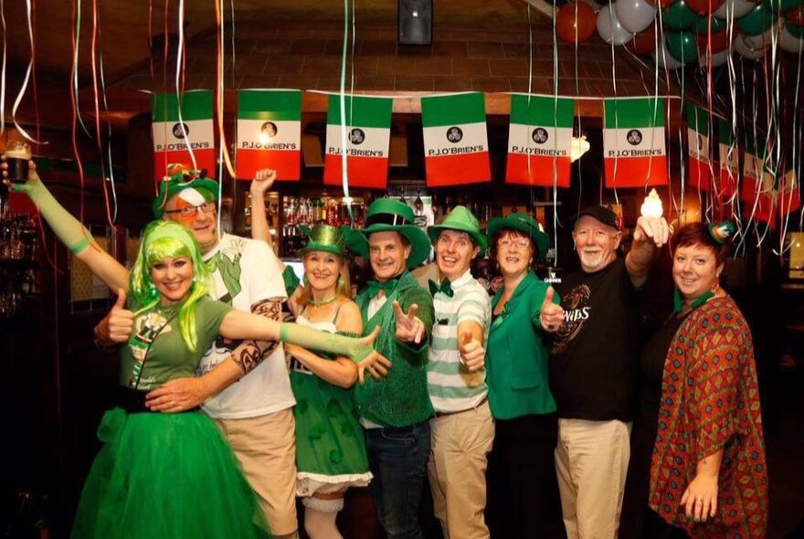 A lively St. Patrick's Day celebration at an Irish pub in Melbourne, featuring patrons dressed in festive green attire standing underneath Irish flags hanging from the ceiling. 