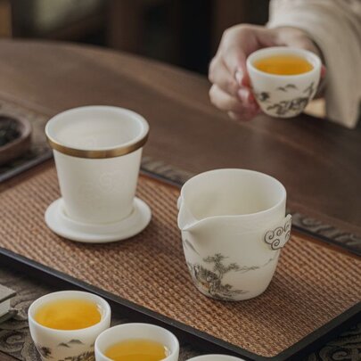 A hand holding a small cup of tea, in the foreground are more small cups of tea and a ceramic pot. 